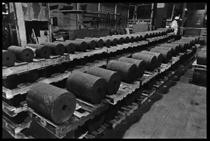 Ingots of Fernald at Ashtabula
These ingots, made at Fernald by remelting uranium metal derbies, have been trucked 300 miles northeast to Ashtabula, Ohio, for further processing. Here they will be submerged in molten salt until they reach a red-hot 1100F. Then thy will be inserted into the Ashtabula uranium metal extrusion press. The sign in the background reads "Caution: Radioactive Materials." 

Reactive Metals, Incorporated, Ashtabula, Ohio. June 19,1984.
Keywords: Fernald Green Salt Plant, Feed Materials Production Center, Fernald, Ohio (FEMP)